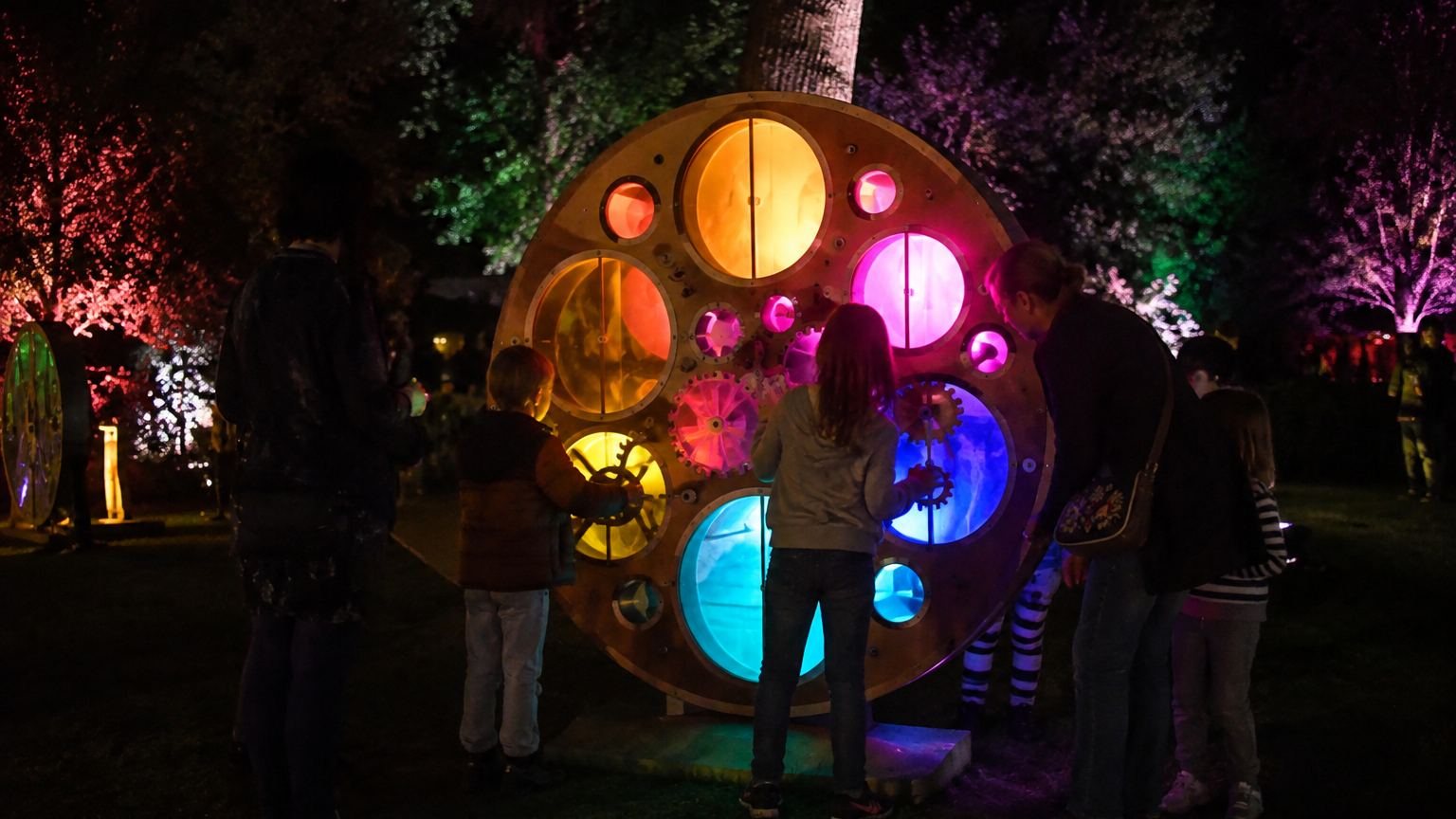Children and adults at night turning the gears of a large wooden wheel set with glowing amber, magenta and blue discs, under trees lit red, green and purple.
